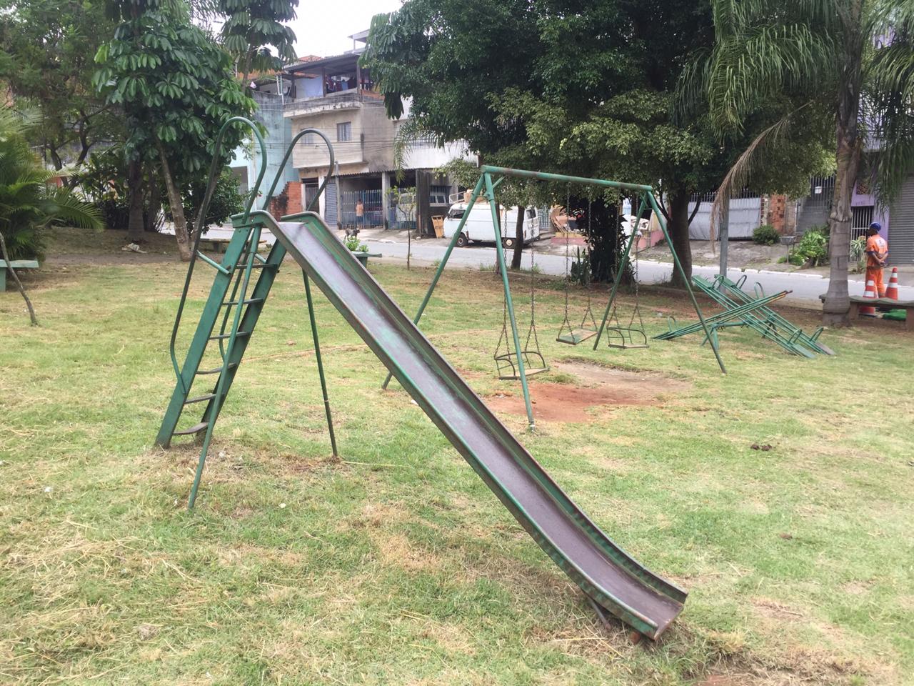 Quatro brinquedos pintados em verde, mas com pintura já desbotada, são destaque na foto. A grama já está cortada e o local limpo. à frente, várias árvores na beira da calçada e no miolo da praça.